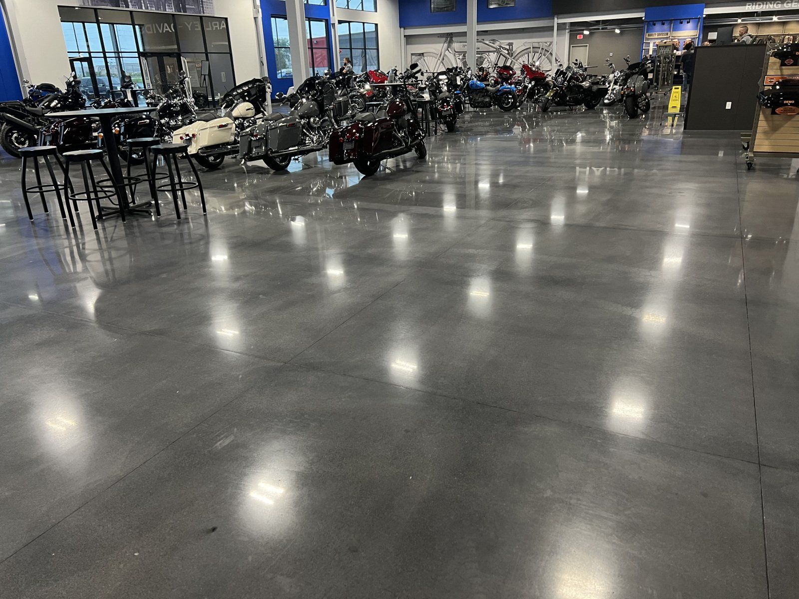 A glossy concrete floor in a dealership showroom displaying rows of motorcycles and a small bistro-style seating area.