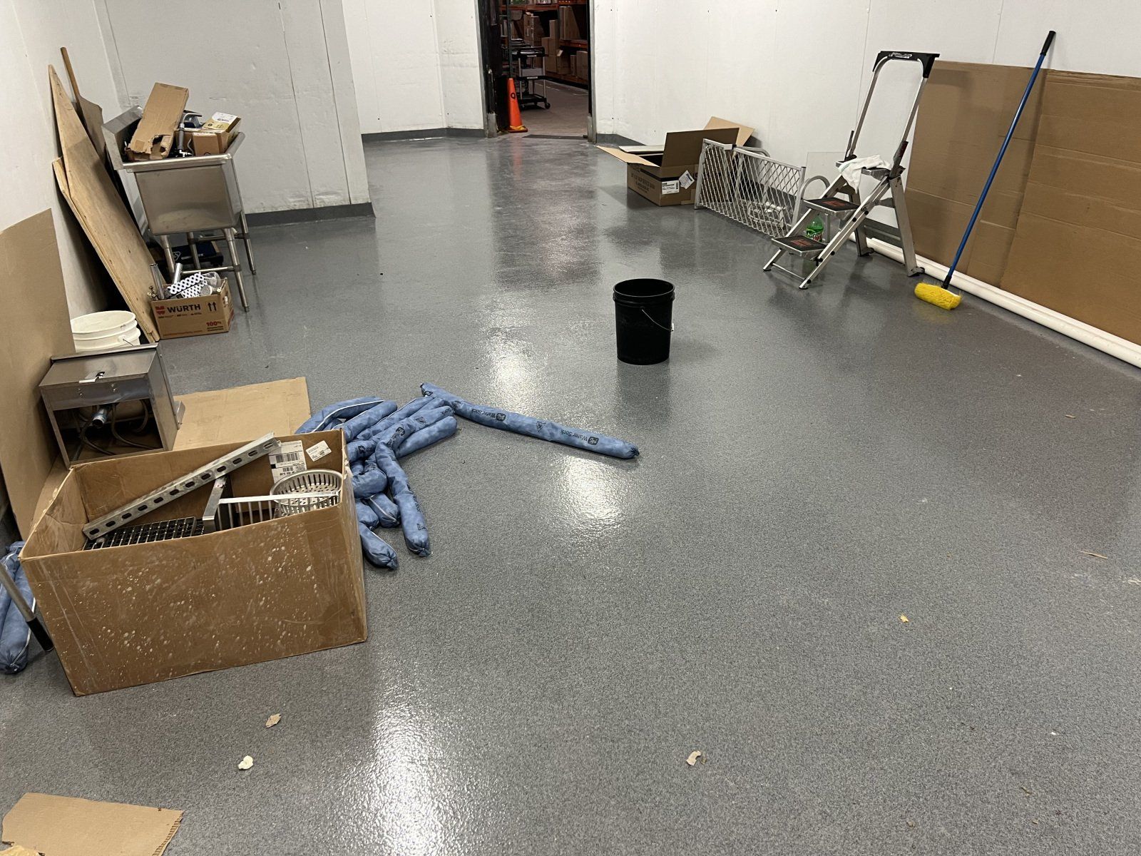 An empty room with gray speckled flooring, featuring cardboard boxes, a black bucket, a stepladder, and a push broom.