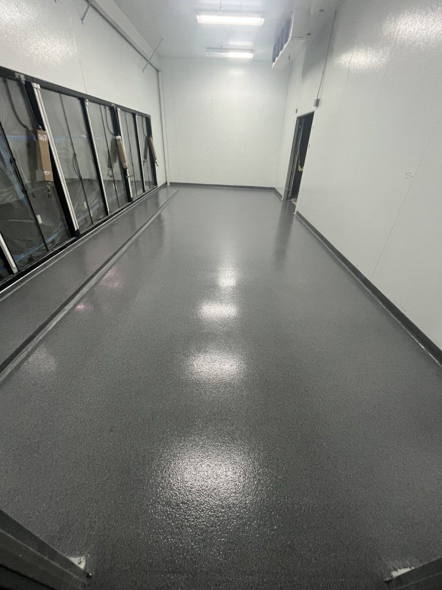 A long, narrow room with white walls, a gray speckled epoxy floor, and a row of glass refrigerator doors on the left.