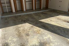 A concrete garage floor with contrasting light and dark patches, showing patches of discoloration and sunlight streaks.