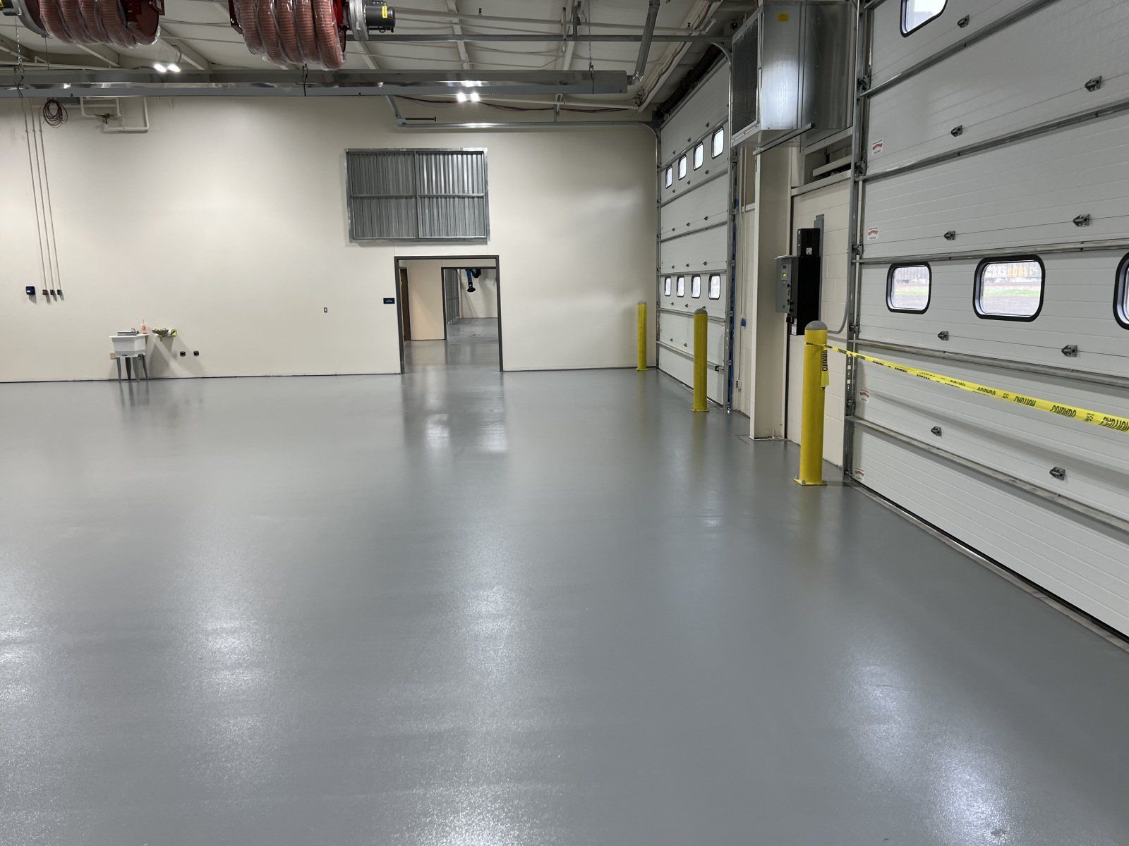 A wide, empty garage with shiny gray epoxy floors, white walls, and a large bay door blocked off by yellow caution tape.