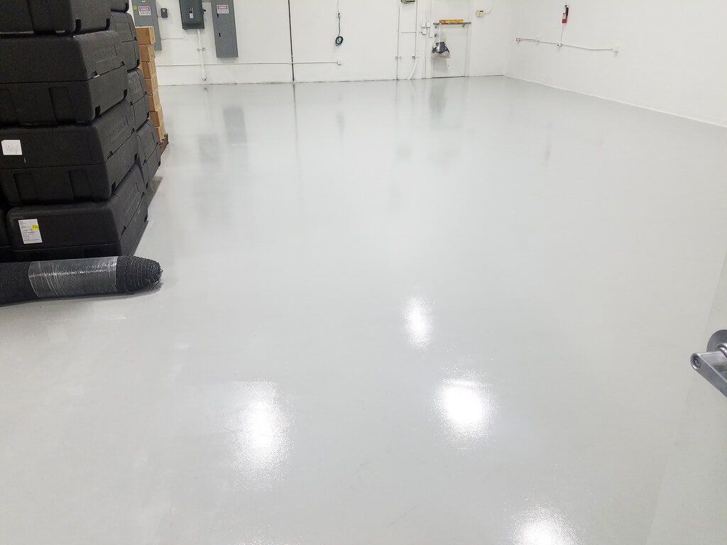 A smooth, high-gloss light gray epoxy floor in a clean, brightly lit industrial storage room with stacked black cases.