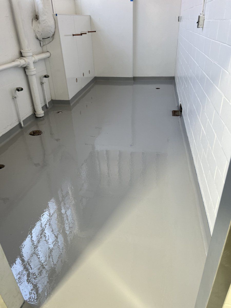 A small, windowless utility room with a smooth, glossy grey epoxy floor, white walls, and exposed plumbing pipes.