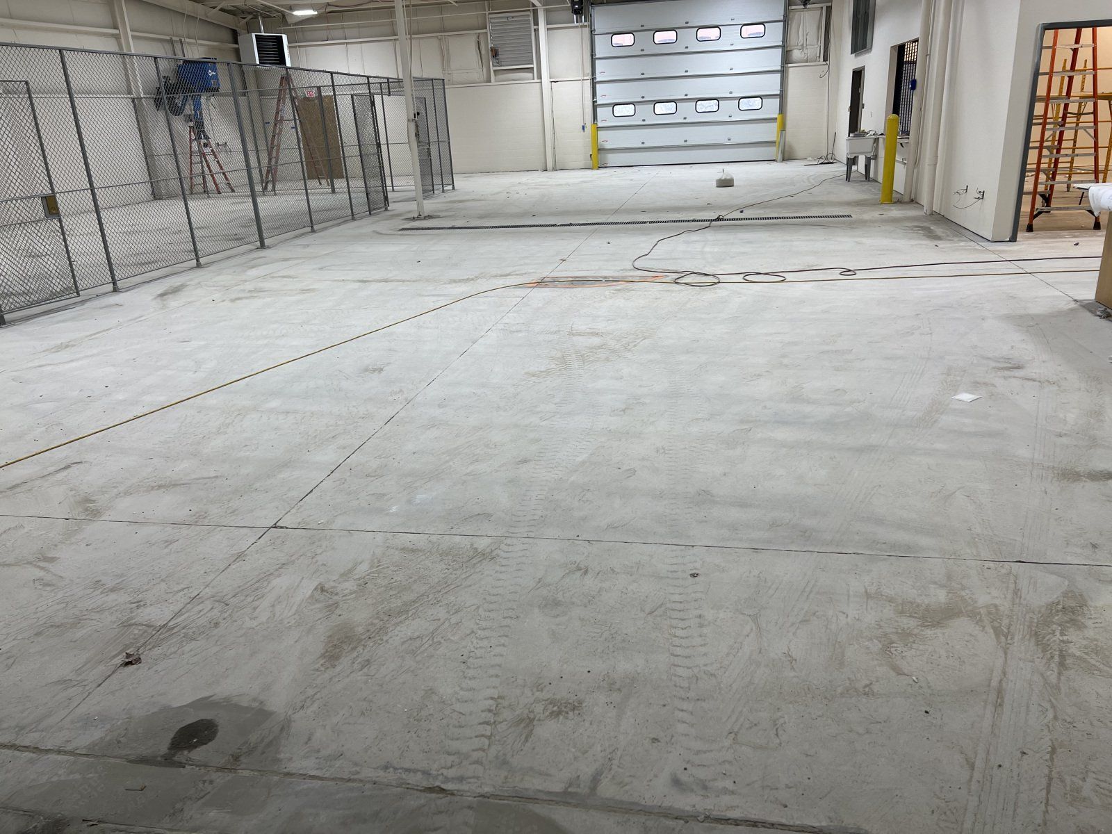 A large, empty concrete warehouse floor with a metal chain-link fence on the left and a large closed bay door in the back.