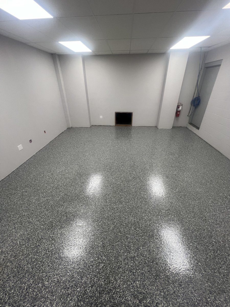 An empty room with light gray walls, a speckled epoxy floor, and overhead fluorescent lighting.