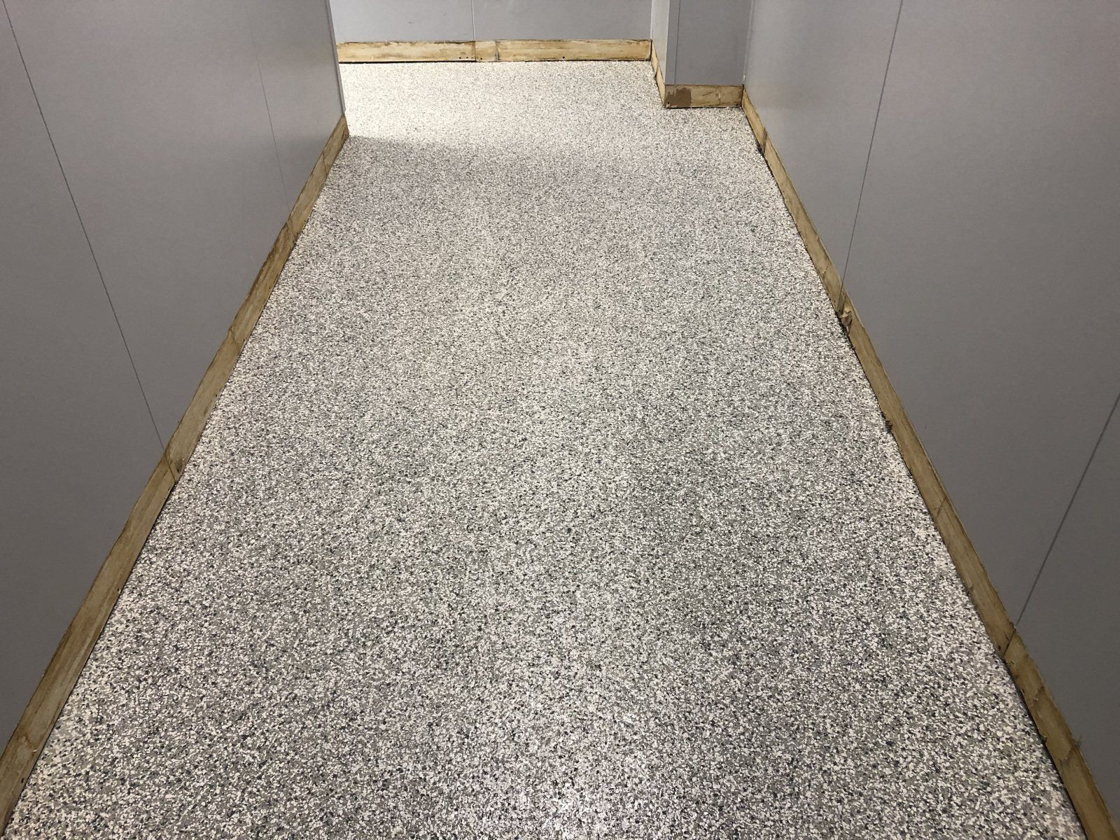 A narrow hallway with grey walls and a textured, speckled stone-look floor lined with wooden trim.
