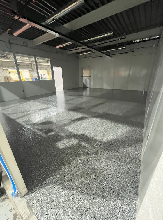 Empty industrial room with gray epoxy floor, exposed ceiling, and a windowed wall.