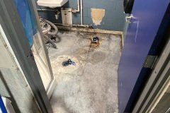 Damaged concrete floor in a utility area with exposed pipes and small holes near a wall.