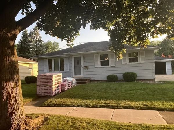 A light gray ranch-style house with stacks of pink building materials on the front lawn under a tree on a sunny day.