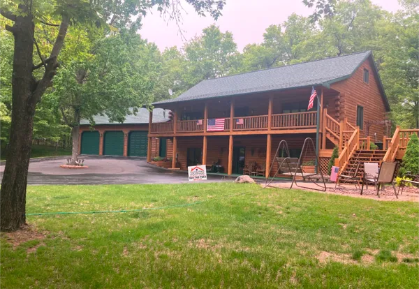 A large two-story log cabin home with a detached three-car garage in a wooded setting, featuring a wrap-around porch.