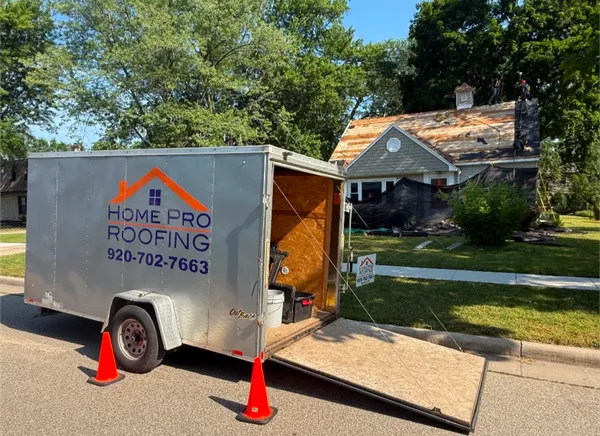 A Home Pro Roofing trailer with an open ramp parked in front of a house undergoing roof repairs on a sunny day.