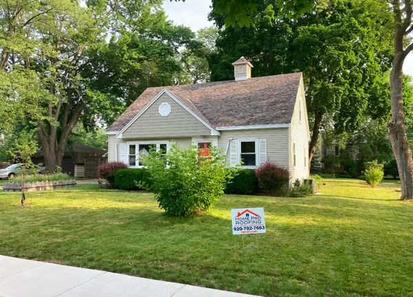A one-story tan house with a steep roof, green lawn, and a real estate sign in the front yard.