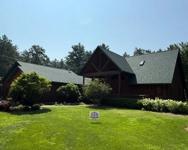 A dark-wood cabin with a green shingled roof, set against a backdrop of trees, with a lawn and a small yard sign.