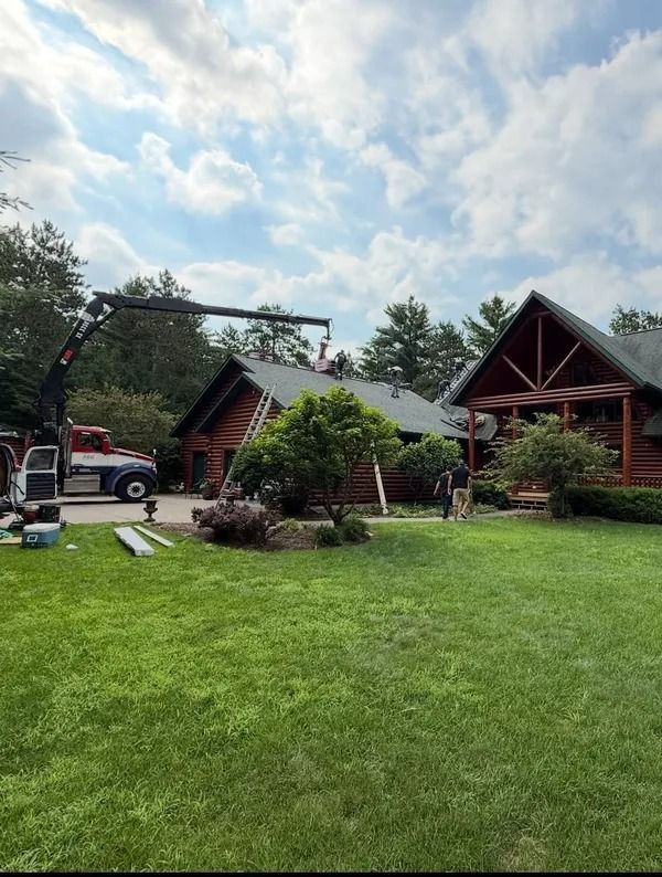 A truck with a crane parked beside a large log cabin, lifting materials to the roof with workers nearby on a sunny day.