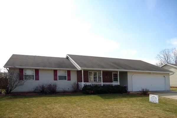 A single-story, light-colored ranch house with a two-car garage, red shutters, and a front porch under a clear blue sky.