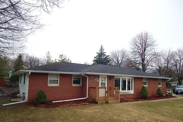 A single-story red brick house with a dark shingled roof, a wooden front deck, and a front-facing bay window.