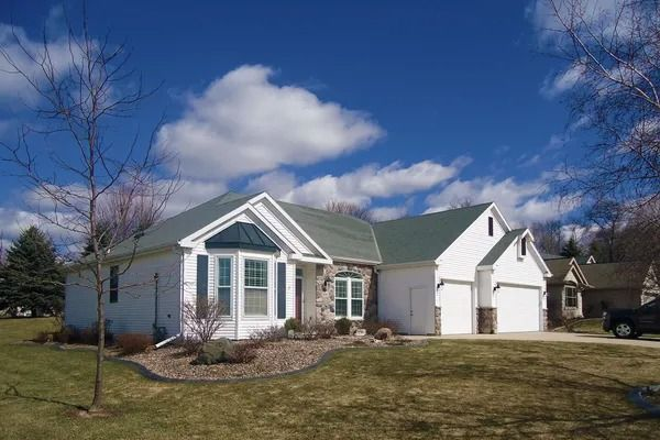 A white, single-story house with green shingles, a stone facade, and a two-car garage sits under a bright, cloudy blue sky.