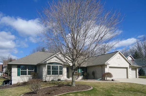 A single-story, light-colored brick house with a green-trimmed window and a bare tree in the front yard under a blue sky.