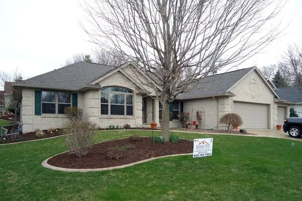 Single-story brick home with a tan exterior, dark shingles, an attached garage, and a landscaped yard with a large tree.