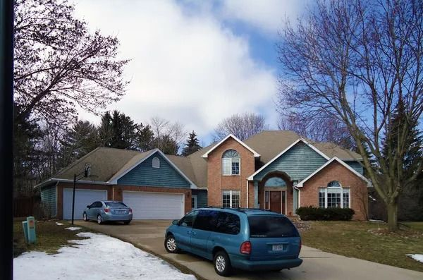 A brick and blue-sided house with a garage and two cars parked in the driveway on a sunny day with patches of snow.