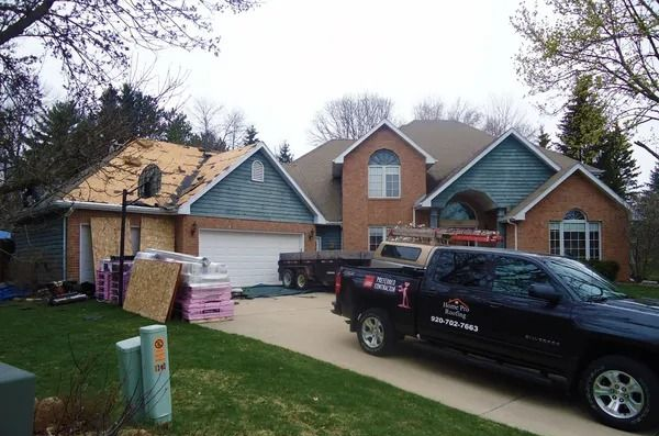 A residential brick house with its garage roof stripped for construction, with supplies and a work truck in the driveway.