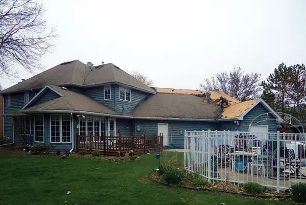 A blue two-story house with a damaged roof showing exposed plywood, a backyard deck, and a white pool fence.