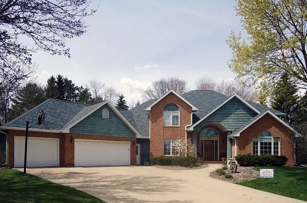 A brick house with a multi-car garage, gray shingled roof, and green siding accents under a partially cloudy sky.