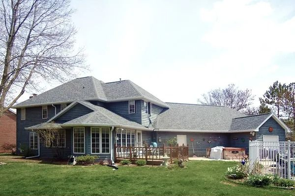 A blue two-story suburban house with a gray shingled roof, a wooden deck, and a spacious green lawn.