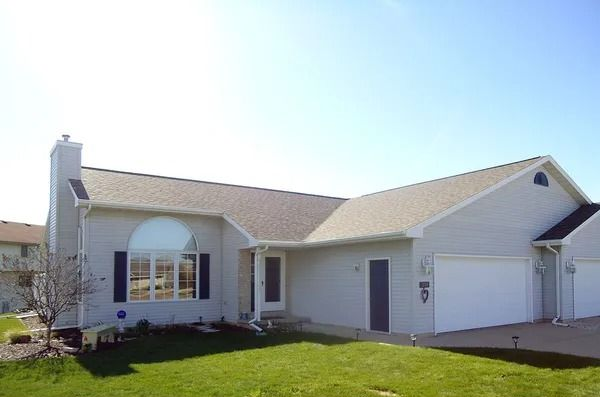 A single-story, light-gray suburban home with a two-car garage, a chimney, and a half-moon window under a clear blue sky.
