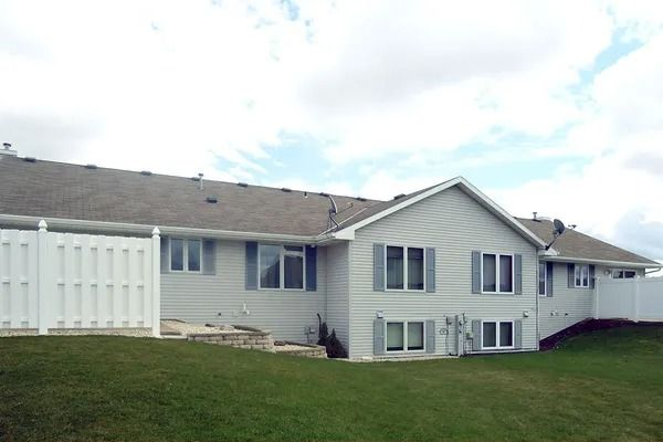 A single-story, light gray house with a white fence on a grassy lot under a partly cloudy sky.