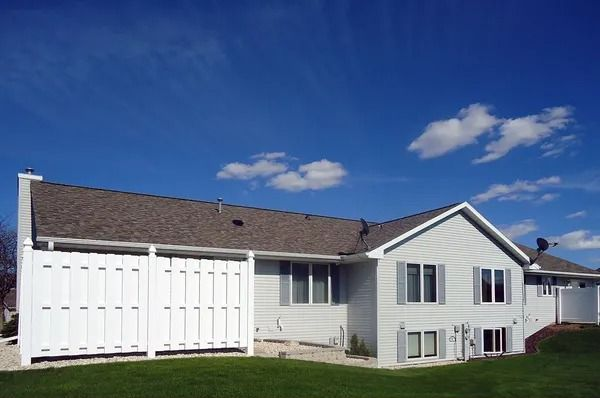 A light-sided suburban house with a large white privacy fence, gray roof, and lawn under a bright blue sky with clouds.
