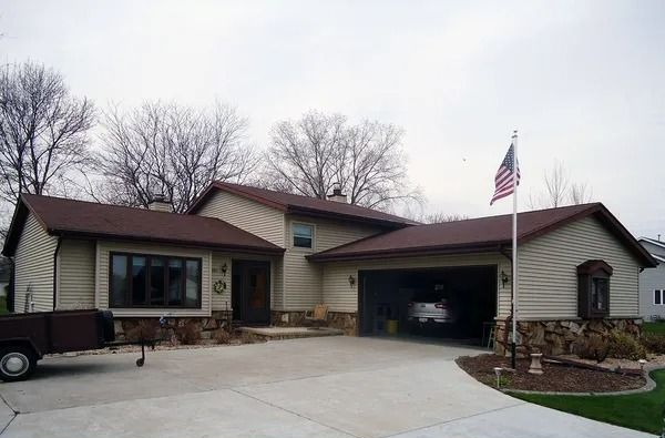A beige suburban house with brown roofing and stone accents features an attached garage, a concrete driveway, and a flag.