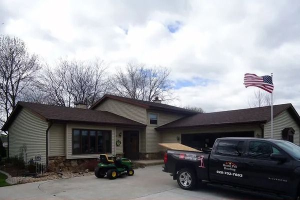 A beige single-story house with a brown roof, a lawn tractor in the driveway, and a work truck with a company logo.