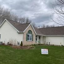 A suburban home with white siding, a stone entryway, and a two-car garage under a cloudy sky, with a sign on the lawn.