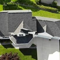 Aerial view of a residential house with a dark gray shingled roof, white trim, and surrounding green lawn.
