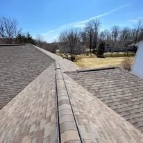 View of a brown shingled roof with a prominent ridge cap, looking out toward trees and a neighboring house under a blue sky.