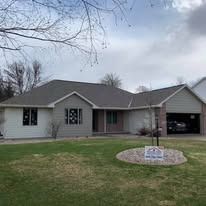 A light gray ranch-style house with a dark roof and garage, surrounded by a lawn and a small circular garden bed.