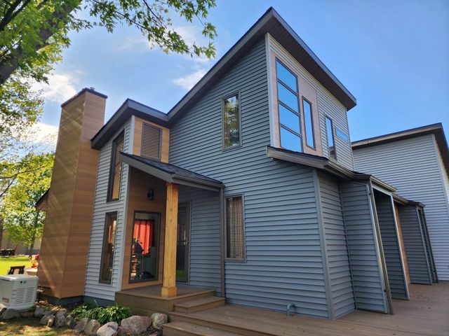 Modern gray house exterior with wood porch, tall windows, and brick chimney under trees.