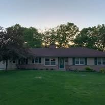 A single-story, light-colored brick house with a dark roof and a green lawn, surrounded by mature trees at dusk.