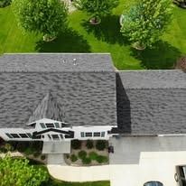 Aerial view of a house with a gray shingled roof, a white exterior, and a surrounding green lawn with trees.