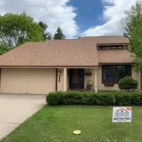 A tan single-story house with a brown shingled roof, a two-car garage, and a green lawn with a roofing company sign.