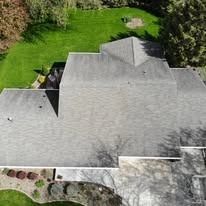 High-angle view of a gray shingled residential roof with surrounding green lawn and trees.