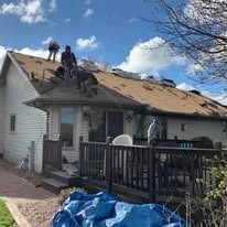 Construction workers repair a shingled roof on a suburban house with a deck in front under a blue, cloudy sky.