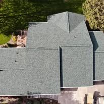 An aerial view of a gray shingled roof with several sections and valleys, surrounded by grass and trees.