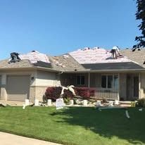 Two roofers installing shingles on the roof of a beige one-story suburban house on a sunny day.