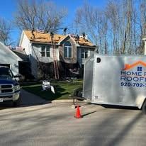 Workers replace a residential roof, with a branded utility trailer parked on a driveway in front of the house.