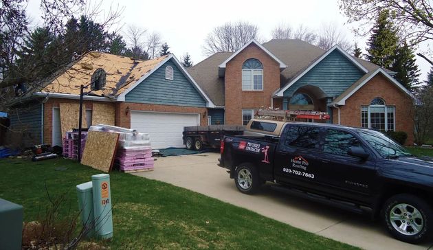 A two-story brick home under renovation with roofing materials and a contractor truck parked in the driveway.
