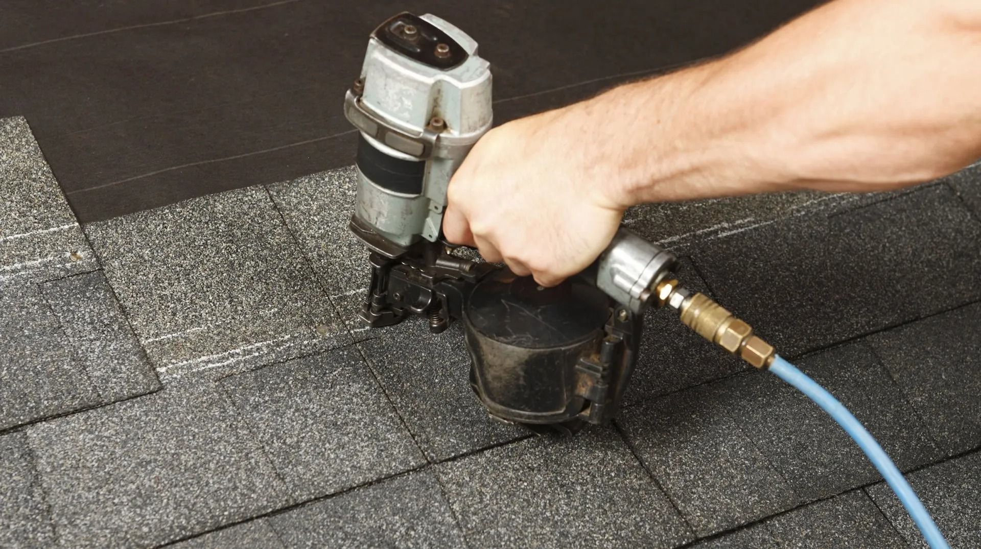 A hand uses a pneumatic nail gun to attach gray shingles to a roof.