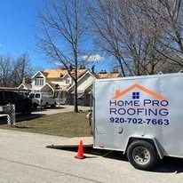 A Home Pro Roofing trailer parked in front of a residential house under construction with roofers working on top.