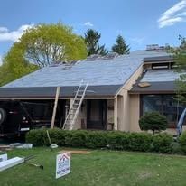 A residential house with a roof under construction, featuring bare plywood, a tall ladder, and a professional yard sign.